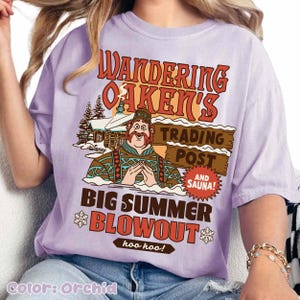 Op de afbeelding: Orchidee-kleurig T-shirt met een afbeelding van een stripfiguur en de tekst "Wandering Oaken's Trading Post and Sauna!" Het shirt heeft ook de tekst "Big Summer Blowout" en "hoo hoo!"