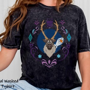 Puede incluir: Camiseta negra lavada con un estampado de Sven y Olaf de Frozen, rodeados de un dise&ntilde;o decorativo morado y turquesa. El texto "Mineral washed Black T-shirt" est&aacute; impreso en la parte inferior de la camiseta.