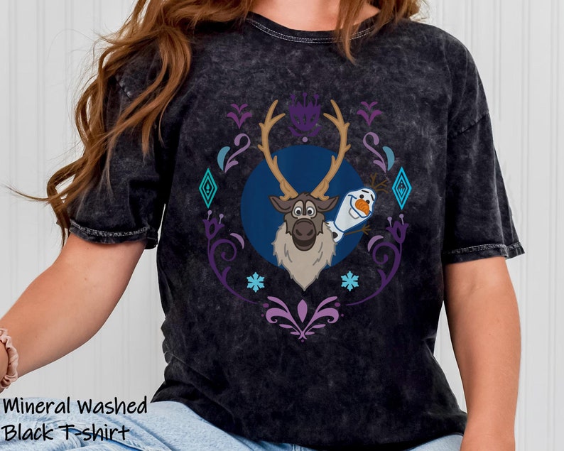 Puede incluir: Camiseta negra lavada con un estampado de Sven y Olaf de Frozen, rodeados de un dise&ntilde;o decorativo morado y turquesa. El texto "Mineral washed Black T-shirt" est&aacute; impreso en la parte inferior de la camiseta.