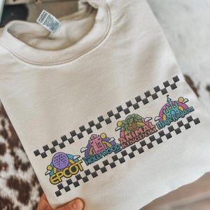 Puede incluir: Una sudadera beige con gr&aacute;ficos coloridos de iconos de parques tem&aacute;ticos, incluyendo Epcot, Hollywood Studios, Animal Kingdom y Magic Kingdom. El dise&ntilde;o est&aacute; bordeado por un patr&oacute;n de cuadros negros y grises. La sudadera est&aacute; doblada.