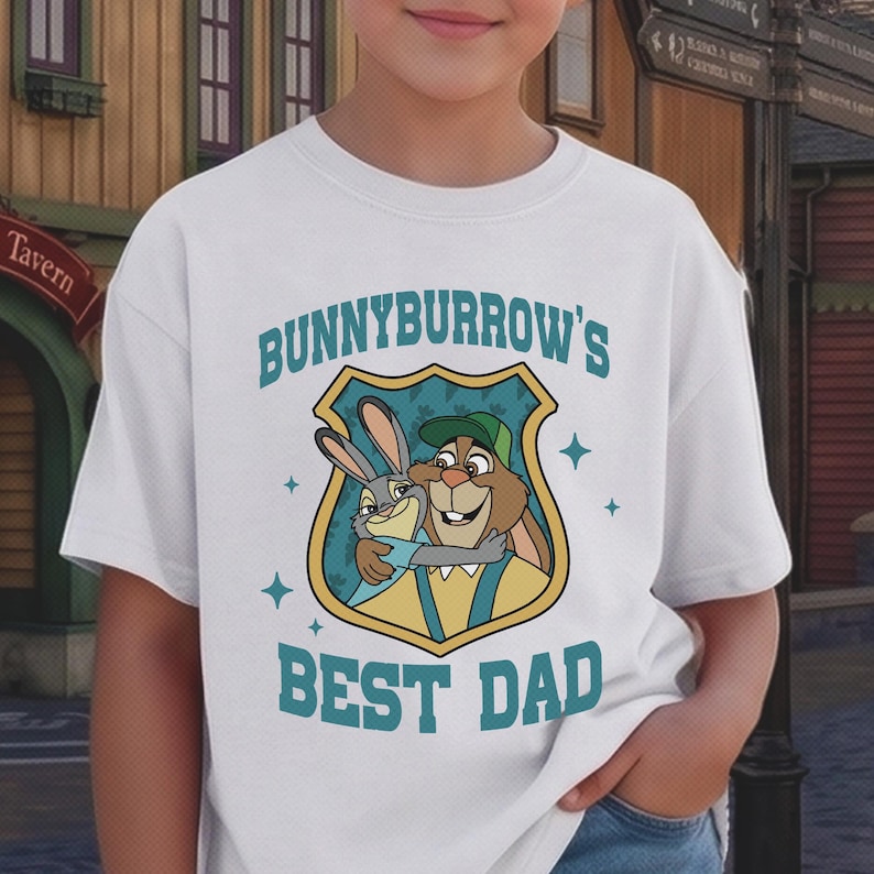 Puede incluir: Camiseta blanca con una imagen de dibujos animados de dos conejos abraz&aacute;ndose dentro de un escudo. El texto "BUNNYBURROW'S BEST DAD" est&aacute; en verde azulado. La camiseta es de cuello redondo y manga corta.