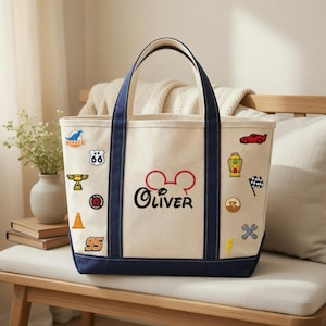 May include: Cream-colored tote bag with navy blue trim and handles. The bag features embroidered patches of a race car, a checkered flag, and other designs. The name "Oliver" is embroidered in black with a red Mickey Mouse head.