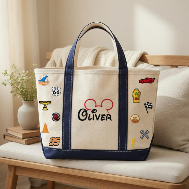 May include: Cream-colored tote bag with navy blue trim and handles. The bag features embroidered patches of a race car, a checkered flag, and other designs. The name "Oliver" is embroidered in black with a red Mickey Mouse head.
