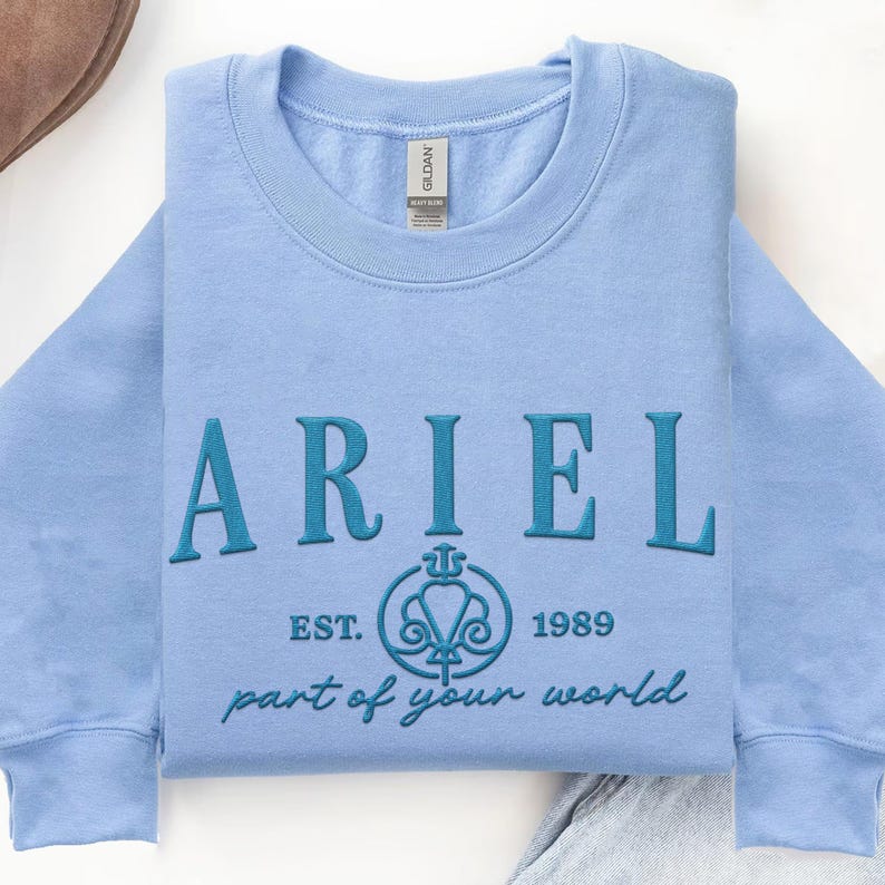 May include: A light blue sweatshirt with the name "ARIEL" embroidered in teal. Below the name is a teal emblem with the text "EST. 1989" and "part of your world" in a script font. The sweatshirt is folded neatly on a white surface.