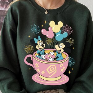 May include: Dark green sweatshirt with a vibrant graphic of Mickey and Minnie Mouse in a pink teacup, surrounded by balloons and fireworks. The design incorporates yellow, pink, and blue, offering a cheerful and festive aesthetic.