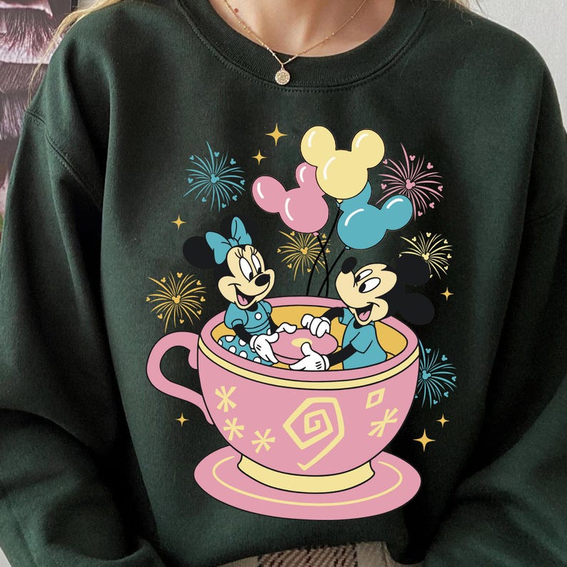 May include: Dark green sweatshirt with a vibrant graphic of Mickey and Minnie Mouse in a pink teacup, surrounded by balloons and fireworks. The design incorporates yellow, pink, and blue, offering a cheerful and festive aesthetic.