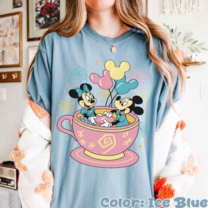 May include: Pale blue t-shirt with a pink teacup design featuring Mickey and Minnie Mouse. The teacup has yellow and pink details. Mickey and Minnie are holding balloons. The text "Color: Ice Blue" is at the bottom of the shirt.
