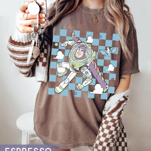 May include: A brown t-shirt with a checkered pattern and a cartoon image of Buzz Lightyear from Toy Story. The image shows Buzz Lightyear flying in space with a spaceship in the background.