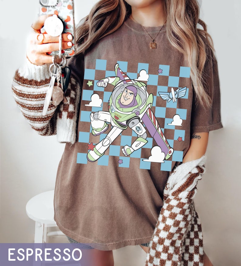 May include: A brown t-shirt with a checkered pattern and a cartoon image of Buzz Lightyear from Toy Story. The image shows Buzz Lightyear flying in space with a spaceship in the background.