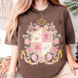 Puede incluir: Camiseta color espresso con un dise&ntilde;o de escudo que presenta una corona, rosas rosas y cintas. El escudo est&aacute; dividido en cuatro secciones con ilustraciones de una l&aacute;mpara, una concha, una flor de loto y un zapato de tac&oacute;n. El color de la camiseta es "Espresso".