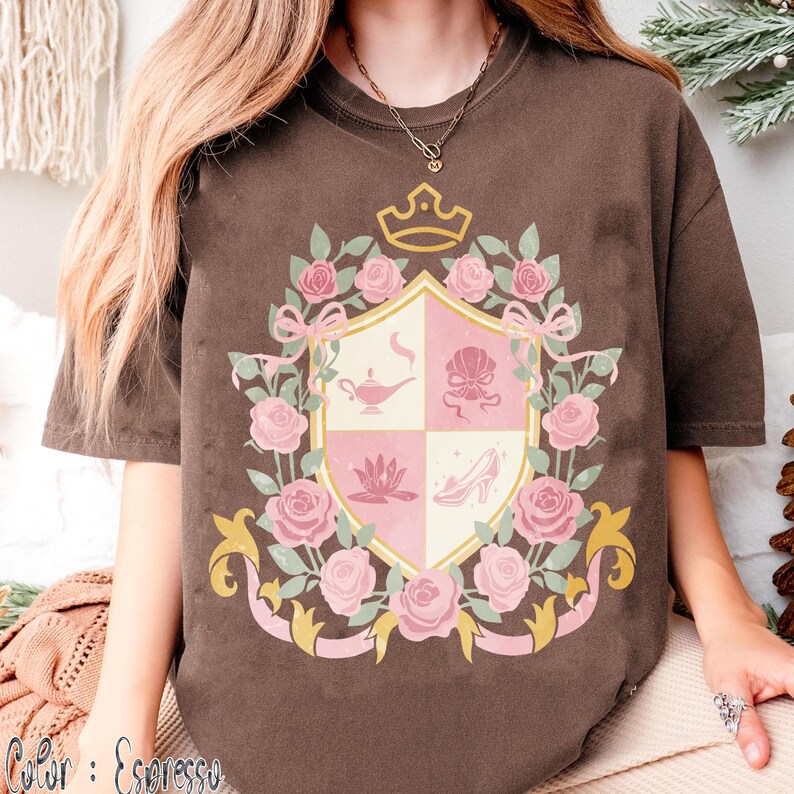 Puede incluir: Camiseta color espresso con un dise&ntilde;o de escudo que presenta una corona, rosas rosas y cintas. El escudo est&aacute; dividido en cuatro secciones con ilustraciones de una l&aacute;mpara, una concha, una flor de loto y un zapato de tac&oacute;n. El color de la camiseta es "Espresso".