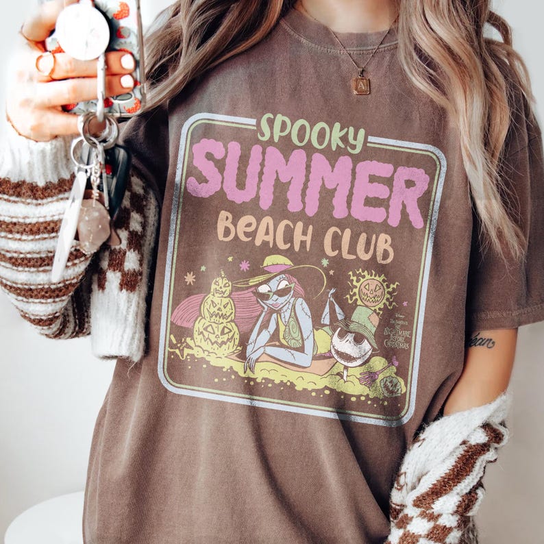 May include: Brown t-shirt with a "Spooky Summer Beach Club" graphic. The design features a cartoon illustration of characters on a beach with a jack-o'-lantern, sun, and palm trees. The text is in pink and green.