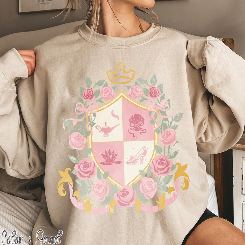 Puede incluir: Sudadera beige con un dise&ntilde;o de escudo con una corona dorada, rosas rosas y un escudo dividido en cuatro secciones. El escudo incluye una l&aacute;mpara, una concha, una flor de loto y un zapato de tac&oacute;n. El texto "Color & Sand" est&aacute; en la parte inferior.
