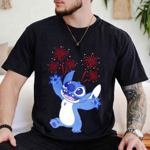 May include: Black t-shirt featuring a blue cartoon character with a white belly and large ears, with red and white fireworks in the background. The character is smiling and has its arms outstretched. The shirt is made of a soft material.