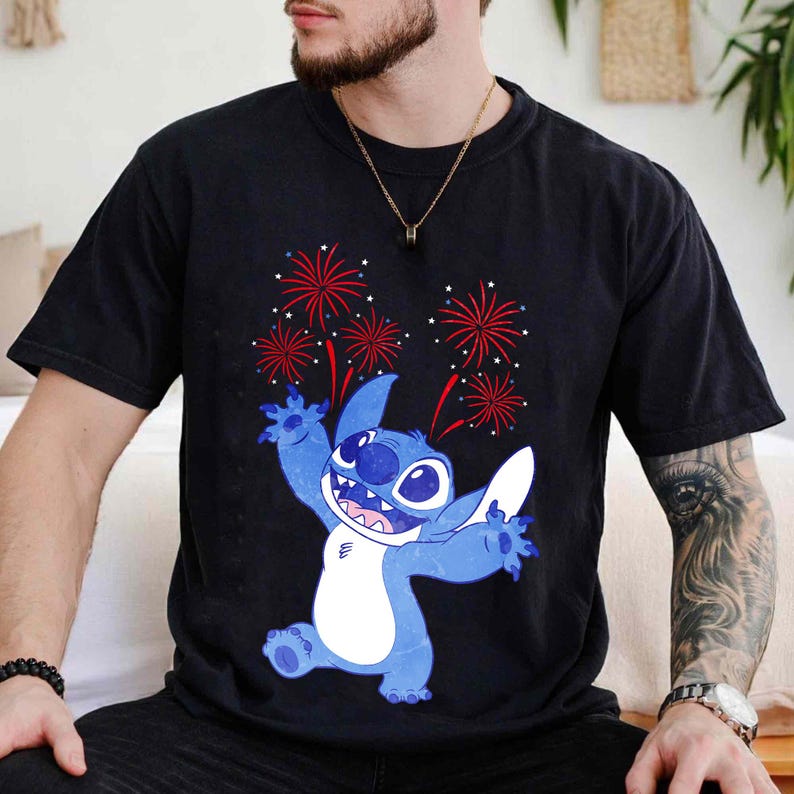 May include: Black t-shirt featuring a blue cartoon character with a white belly and large ears, with red and white fireworks in the background. The character is smiling and has its arms outstretched. The shirt is made of a soft material.