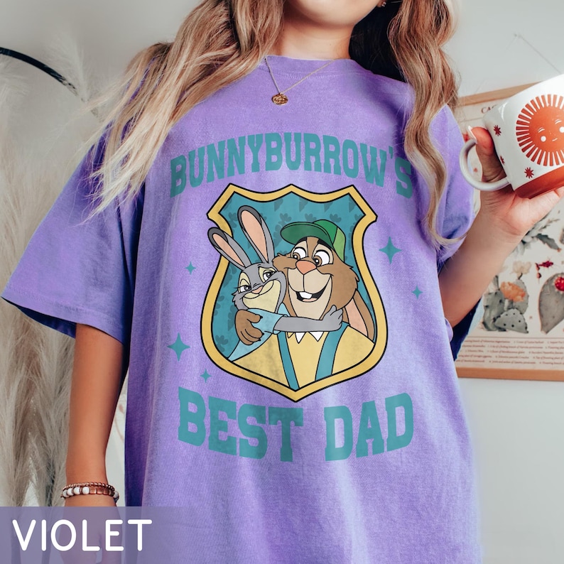 Puede incluir: Camiseta violeta con el texto "BUNNYBURROW'S BEST DAD" en color turquesa. La camiseta presenta una imagen de dibujos animados de dos conejos abraz&aacute;ndose dentro de un escudo. La camiseta es de color violeta liso.