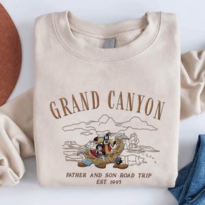 May include: Beige sweatshirt with embroidered text "GRAND CANYON" and a cartoon illustration of two characters on a road trip. The text "FATHER AND SON ROAD TRIP EST. 1995" is also embroidered on the sweatshirt.