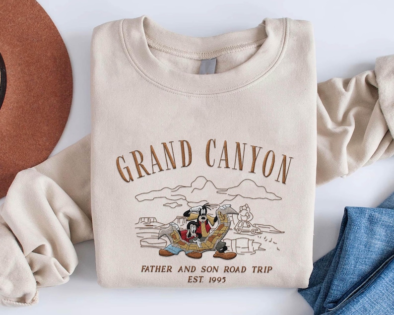 May include: Beige sweatshirt with embroidered text "GRAND CANYON" and a cartoon illustration of two characters on a road trip. The text "FATHER AND SON ROAD TRIP EST. 1995" is also embroidered on the sweatshirt.
