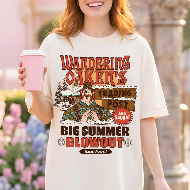 Op de afbeelding: Cr&egrave;mekleurig T-shirt met de tekst "Wandering Oaken's Trading Post and Sauna! Big Summer Blowout" in rood en bruin. De afbeelding toont een stripfiguur en een besneeuwde hut. Een roze koffiekopje wordt in de hand gehouden.