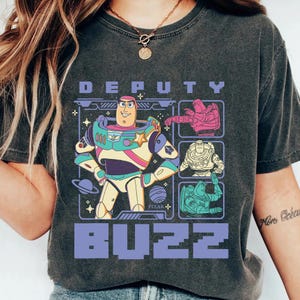 May include: Dark gray t-shirt featuring a colorful graphic of Buzz Lightyear from the movie Toy Story. The design includes the words "DEPUTY BUZZ" in purple block letters, along with images of Buzz in different poses. The Pixar logo is also visible.