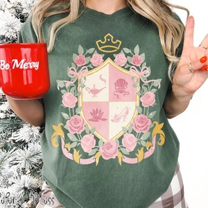Puede incluir: Camiseta verde con un dise&ntilde;o de escudo con rosas rosas, una corona e iconos tem&aacute;ticos de cuentos de hadas. La camiseta tiene un corte holgado. Una taza roja con "Be Merry" est&aacute; en la mano izquierda.