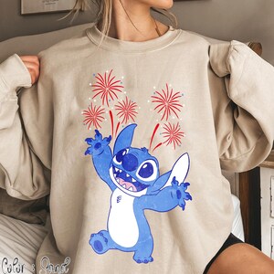 May include: Beige sweatshirt featuring a blue cartoon character with white accents, arms raised in a celebratory pose. Red and white fireworks burst above the character. The sweatshirt has a relaxed fit.