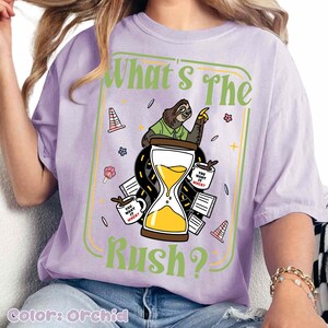 Puede incluir: Camiseta color orqu&iacute;dea con un gr&aacute;fico que presenta un perezoso, un reloj de arena y el texto "What's The Rush?". El dise&ntilde;o incluye tazas de caf&eacute;, elementos de carretera y la frase "You Want It When?" en las tazas. La camiseta es de corte holgado.