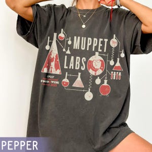 May include: Pepper-colored t-shirt with a graphic design featuring the text "Muppet Labs" and laboratory equipment illustrations in red and white. The shirt also includes text for the Epcot International Food & Wine Festival 2025.