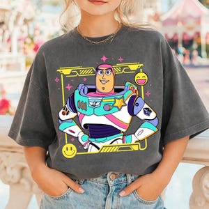 May include: Dark gray t-shirt featuring a colorful cartoon graphic of Buzz Lightyear in his space suit. The design includes a yellow frame, pink stars, and smiley faces. The shirt is a relaxed fit.