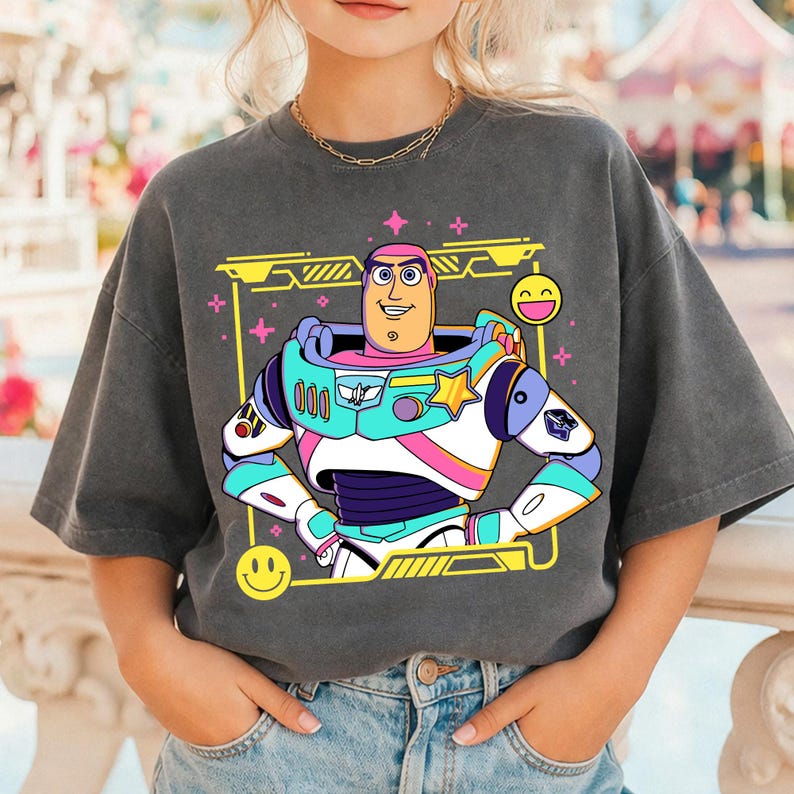 May include: Dark gray t-shirt featuring a colorful cartoon graphic of Buzz Lightyear in his space suit. The design includes a yellow frame, pink stars, and smiley faces. The shirt is a relaxed fit.