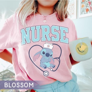 May include: Pink t-shirt with the word "NURSE" in light blue block letters. A cartoon character wearing a nurse's cap is inside a stethoscope-shaped design. The shirt also has the word "BLOSSOM" at the bottom.