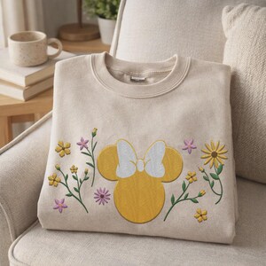 Pu&ograve; includere: Una felpa girocollo beige con una silhouette ricamata gialla di una testa di topo con un fiocco bianco, circondata da piccoli fiori ricamati nelle tonalit&agrave; del giallo, arancione e viola sulla parte anteriore del capo.