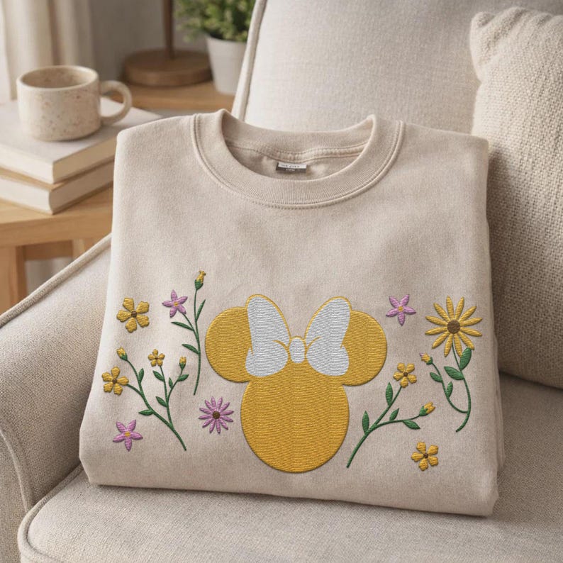 Pu&ograve; includere: Una felpa girocollo beige con una silhouette ricamata gialla di una testa di topo con un fiocco bianco, circondata da piccoli fiori ricamati nelle tonalit&agrave; del giallo, arancione e viola sulla parte anteriore del capo.
