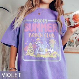 May include: Violet t-shirt with a graphic that reads "Spooky Summer Beach Club." The design features cartoon characters, a pumpkin, and a sun. The shirt is a relaxed fit.