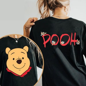May include: Black t-shirt featuring a cartoon image of Winnie the Pooh on the front. The back of the shirt displays the word "POOH" in red, with bees incorporated into the design. A casual, comfortable style.