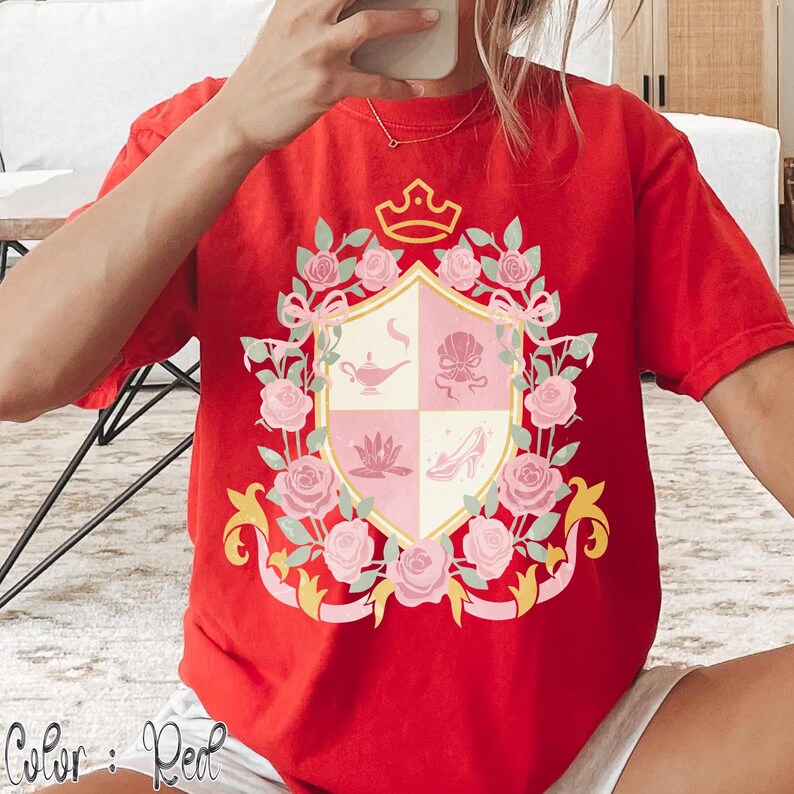 Puede incluir: Camiseta roja con un dise&ntilde;o de escudo que incluye una corona, rosas y un escudo dividido en cuatro secciones. El escudo contiene im&aacute;genes de una l&aacute;mpara, una concha, una flor de loto y un tac&oacute;n alto. El texto "Color: Red" est&aacute; en la parte inferior.