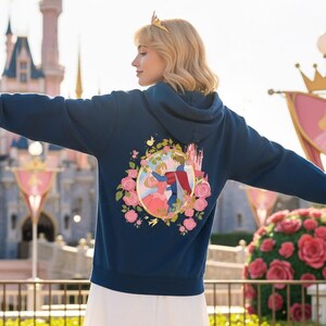 May include: A navy blue hoodie featuring a colorful graphic of a couple dancing in a floral frame, with a castle in the background. The hoodie has a hood and long sleeves. The design is reminiscent of a fairytale.