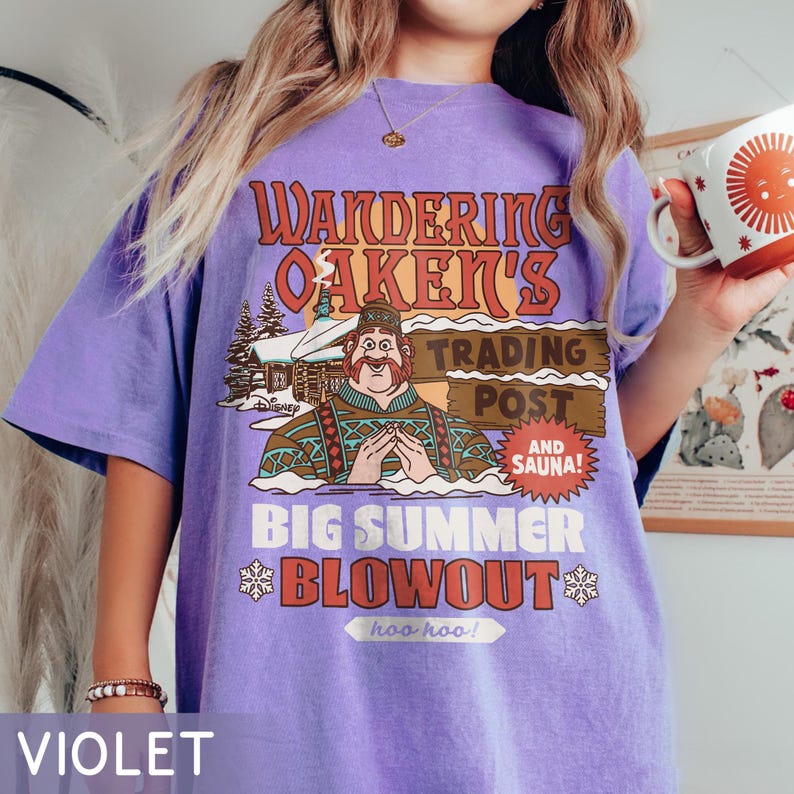 Op de afbeelding: Violet t-shirt met een afbeelding met de tekst "Wandering Oaken's Trading Post and Sauna!" en een cartoonafbeelding van een man. Het ontwerp bevat sneeuwvlokken en de woorden "Big Summer Blowout".