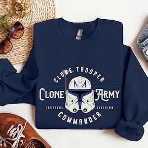 Op de afbeelding: Navyblauw sweatshirt met een witte en blauwe Star Wars Clone Trooper helm afbeelding. De tekst "Clone Trooper Clone Army Tactical Division Commander" is rond de helm gedrukt.
