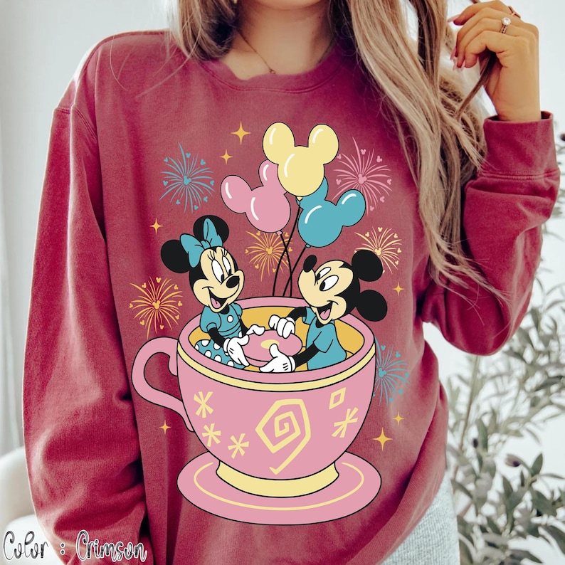 May include: Crimson sweatshirt with a graphic of Mickey and Minnie Mouse in a pink teacup, balloons, and fireworks. The design includes the text "Color: Crimson". The characters are wearing blue outfits. The sweatshirt is a deep red colour.