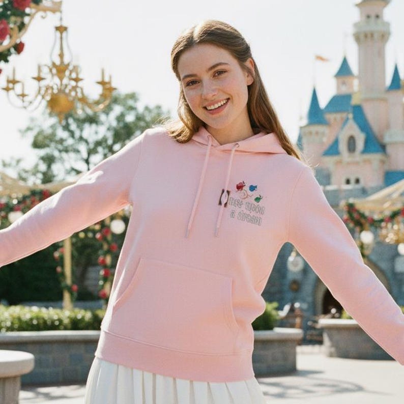 May include: A light pink hoodie with a front pocket and a drawstring hood. The hoodie features embroidered floral and text details that read "Once upon a dream". The background includes a castle and outdoor scenery.