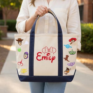 K&ouml;nnte beinhalten: Cremefarbene Canvas-Tragetasche mit marineblauen Akzenten und Griffen. Die Tasche hat aufgestickte Charakteraufn&auml;her und den Namen "Emily" in roter Schrift. Ein lustiges und personalisiertes Accessoire.