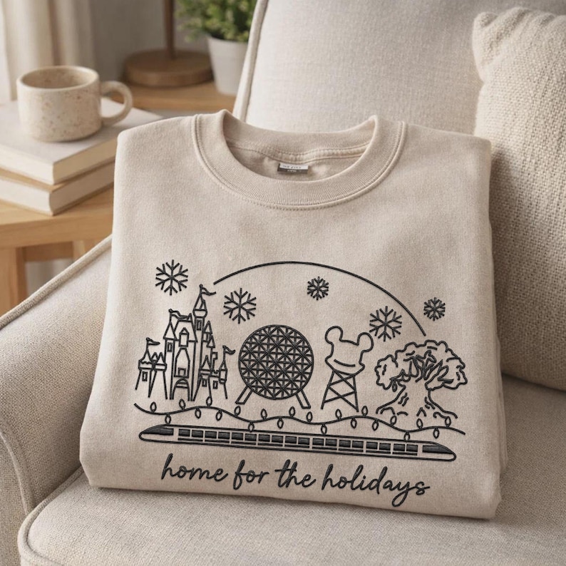May include: A beige crewneck sweatshirt featuring a black line art graphic of a castle, a geodesic sphere, a mouse-ear shaped structure, and a large tree, all under a string of lights and snowflakes with the text "home for the holidays" below.