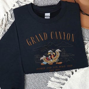 May include: Navy blue sweatshirt with embroidered text "Grand Canyon" and a cartoon illustration of characters on a road trip. The text "Father and Son Road Trip EST. 1995" is also embroidered on the sweatshirt.