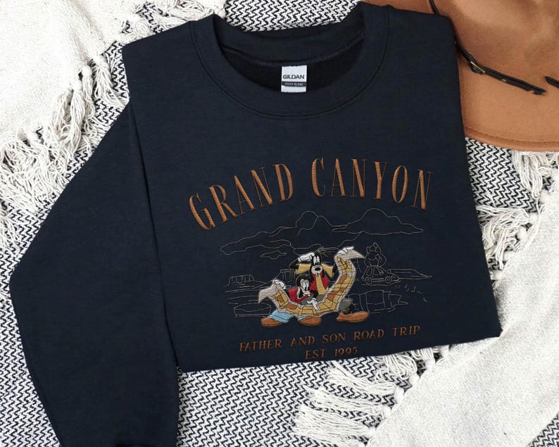 May include: Navy blue sweatshirt with embroidered text "Grand Canyon" and a cartoon illustration of characters on a road trip. The text "Father and Son Road Trip EST. 1995" is also embroidered on the sweatshirt.