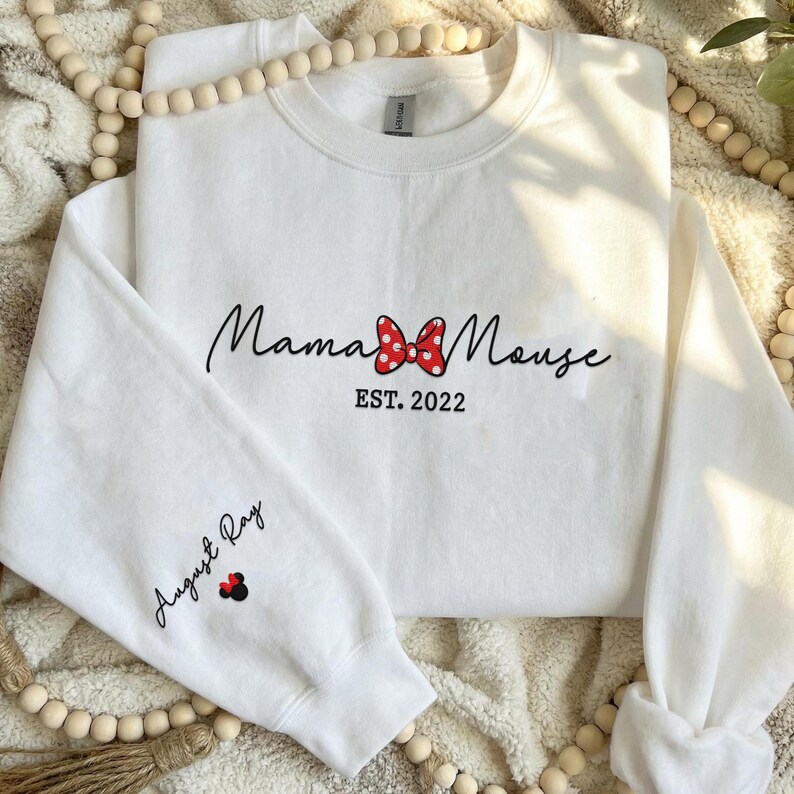 May include: A white long-sleeve sweatshirt featuring the black text Mama Mouse with a red and white polka dot bow graphic in the center and the text EST. 2022 below it. The left sleeve displays the name August Ray in black script with a small bow.