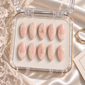 May include: A set of ten oval-shaped press-on nails in a soft blush pink color, featuring intricate white lace patterns and small white pearls. Two of the nails are accented with gold-colored letters A and J, all displayed in a clear plastic case.