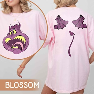 Pode incluir: Camiseta rosa claro com um personagem de desenho animado na frente e atr&aacute;s. A frente apresenta um personagem roxo com dentes amarelos e uma l&iacute;ngua rosa. A parte de tr&aacute;s apresenta asas de morcego e uma cauda roxas. A palavra "BLOSSOM" est&aacute; na parte inferior.