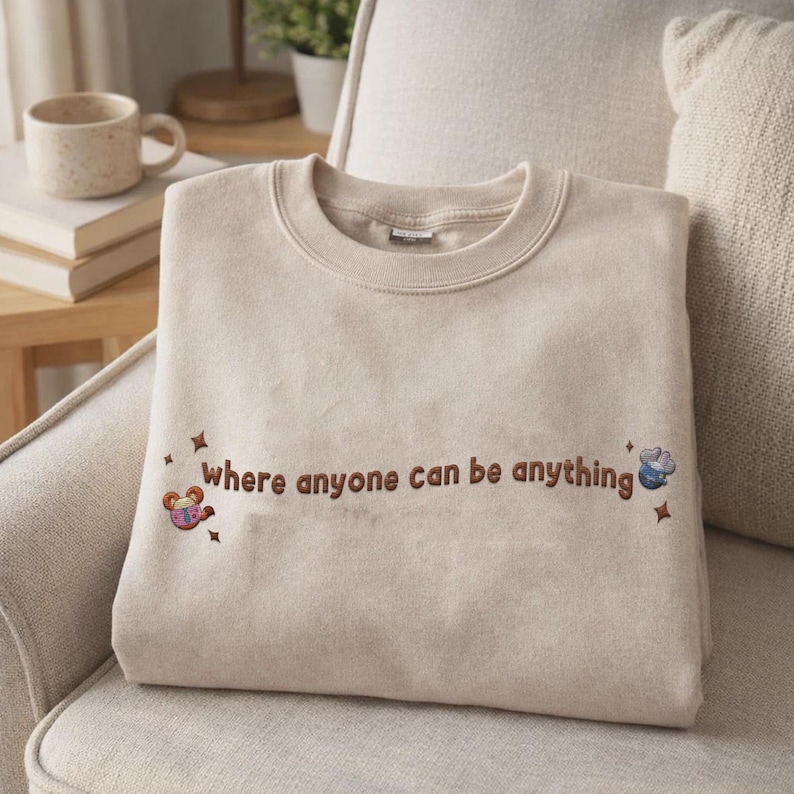 K&ouml;nnte beinhalten: Ein beigefarbenes Sweatshirt mit Rundhalsausschnitt und braun gesticktem Text "where anyone can be anything". Das Design enth&auml;lt kleine gestickte Sternakzente und zwei farbenfrohe Cartoon-Charakter-Symbole auf der linken und rechten Seite.