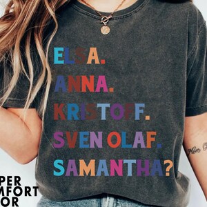 以下が含まれることがあります： 「エルサ」、「アナ」、「クリストフ」、「スヴェンオラフ」、そして「サマンサ？」という名前がカラフルなブロックレターでプリントされた、濃いグレーのTシャツ。「Pepper Comfort Color」の文字が下部にプリントされています。